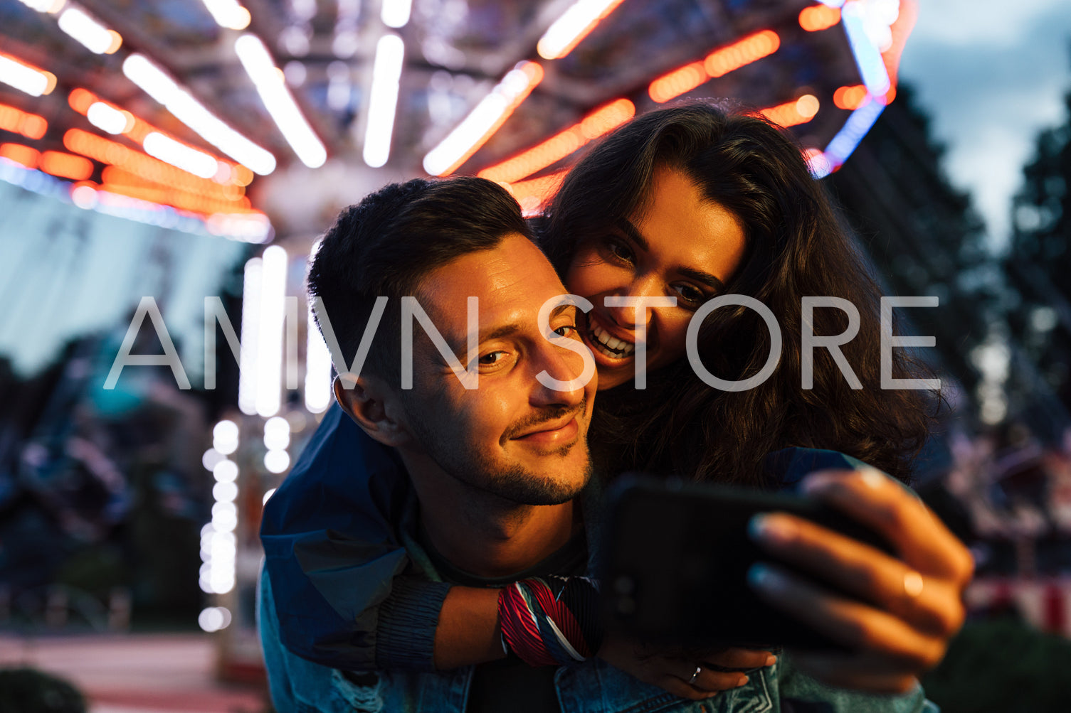 Happy female piggyback her boyfriend and takes a selfie against the carousel. Young couple having fun in an amusement park during the festival.