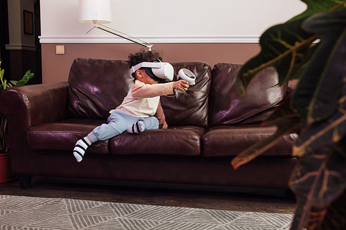 Little boy wearing VR glasses while sitting on sofa. Kid with virtual reality goggles and controller playing a video game at home.