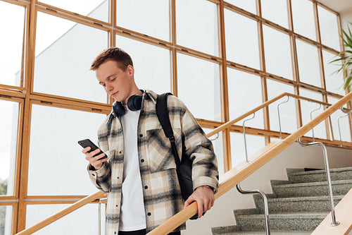 Male student in casuals holds smartphone while standing on stairs