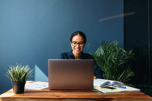 Smiling entrepreneur sitting at home working on laptop computer with documents on the table