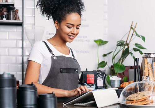 Barista in apron typing on digital tablet. Cheerful woman with curly hair working as a barista in a coffee shop using a digital tablet.