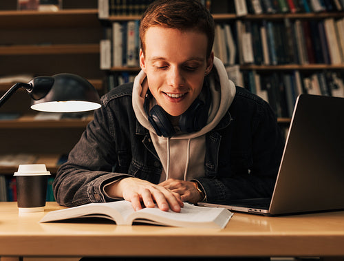 Smiling teenager reading from book. Student with laptop and a book sitting at desk in library.