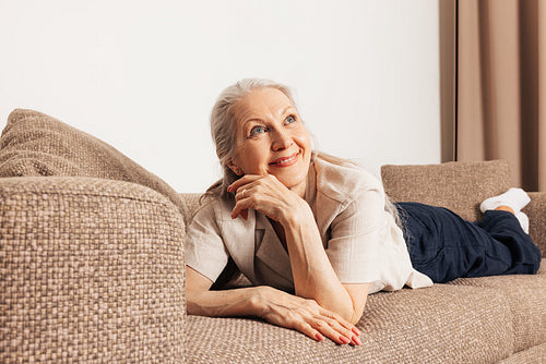 Dreaming senior woman on a sofa. Aged female looking away and smiling in the living room.