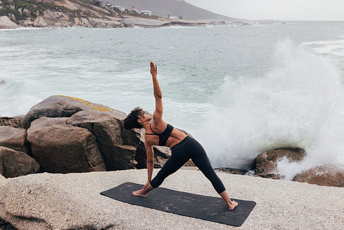 Young female doing Triangle yoga pose against ocean and waves. Full length of a woman practicing stretching exercises at sunset.