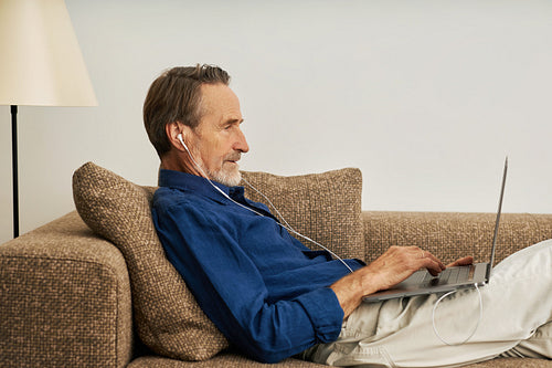 Side view of a handsome senior man lying on sofa typing on laptop