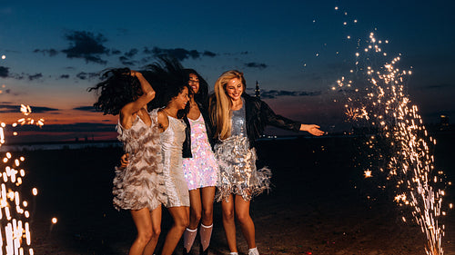 Group of happy girlfriends dancing between fireworks at night. Young stylish women celebrating at sunset.