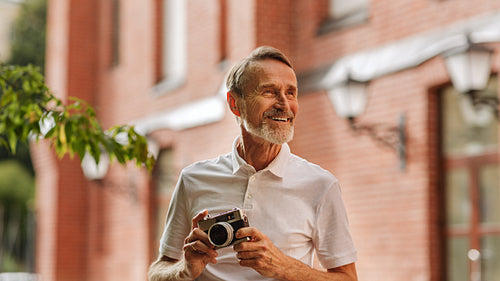 Smiling tourist looking away. Senior man standing outdoors with a film camera outdoors.