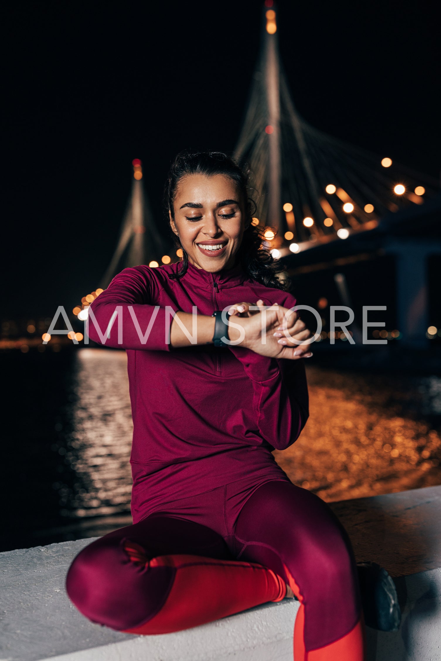 Smiling woman in sportswear sitting at night against bridge checking activity tracker