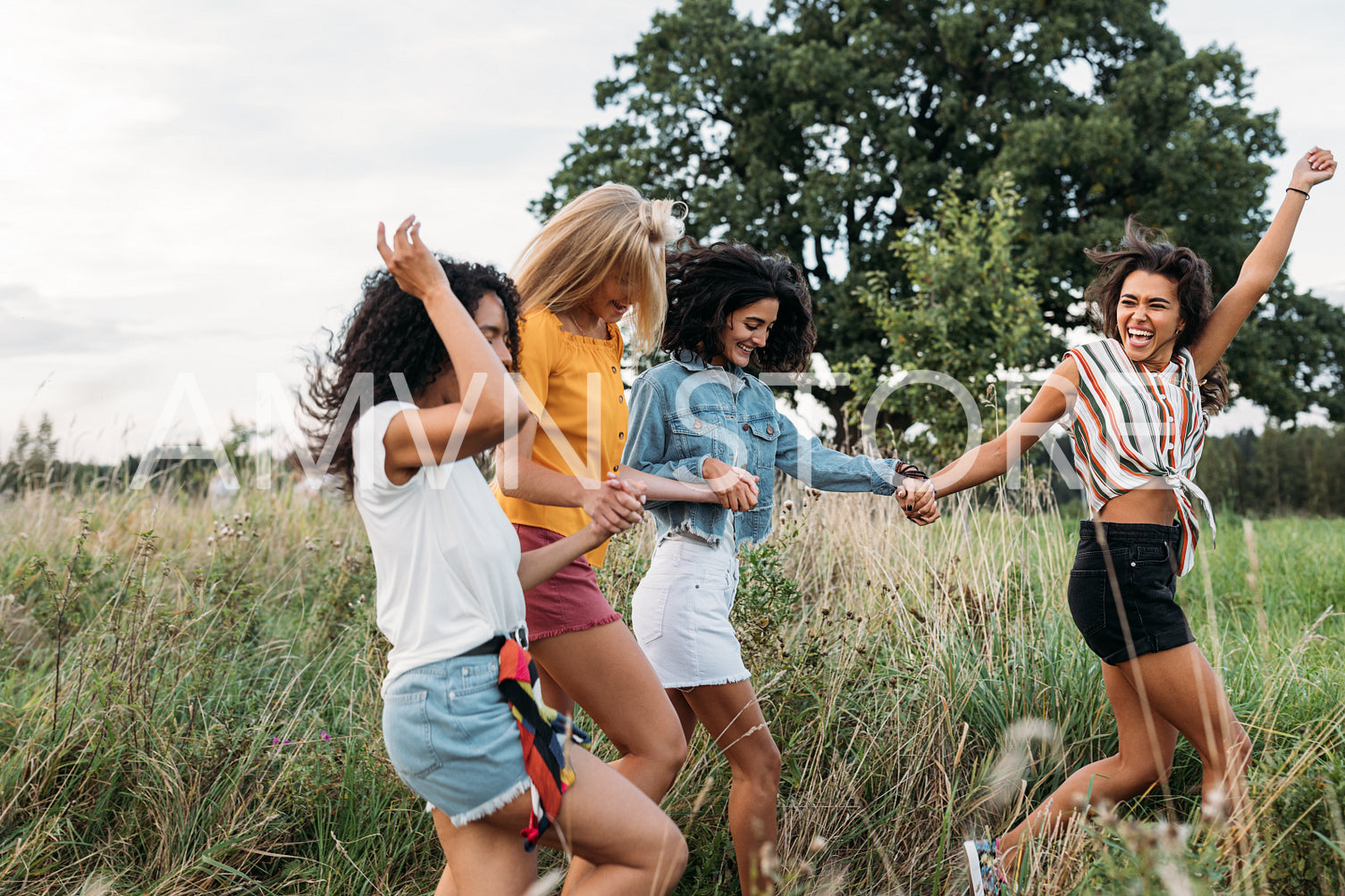 Laughing woman runs in front of her friends with a raised hand. Group of four young females on summer vacation.