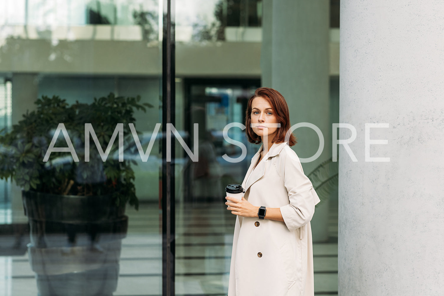 Stylish businesswoman in a coat holding a cup of coffee standing against a glass building