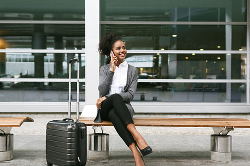 Smiling businesswoman sitting on bench and talking on mobile phone at airport terminal