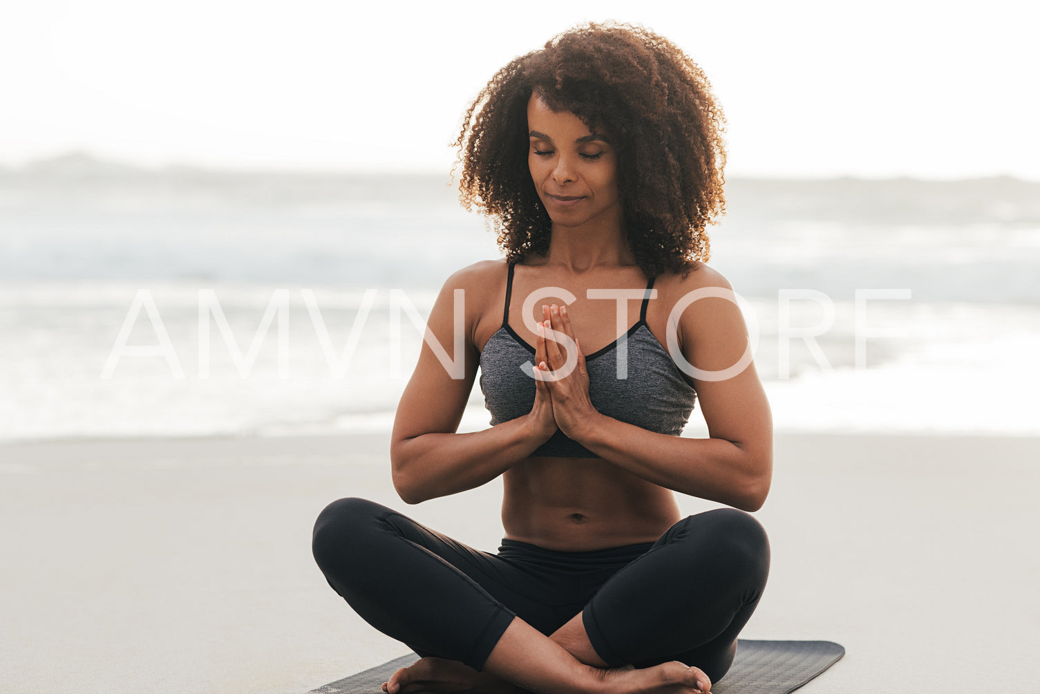 Slim female with closed eyes folded hands while sitting in Sukhasana pose at sunset