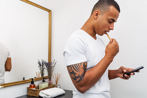 Young man in bathroom brushing his teeth while texting on mobile phone