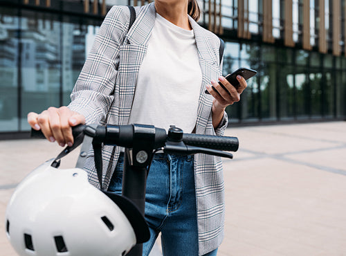 Unrecognizable businesswoman standing in front of an office building with electrical scooter and holding a smartphone