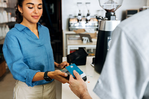 Female coffee shop owner holding a POS terminal while unrecognizable customer paying cashless