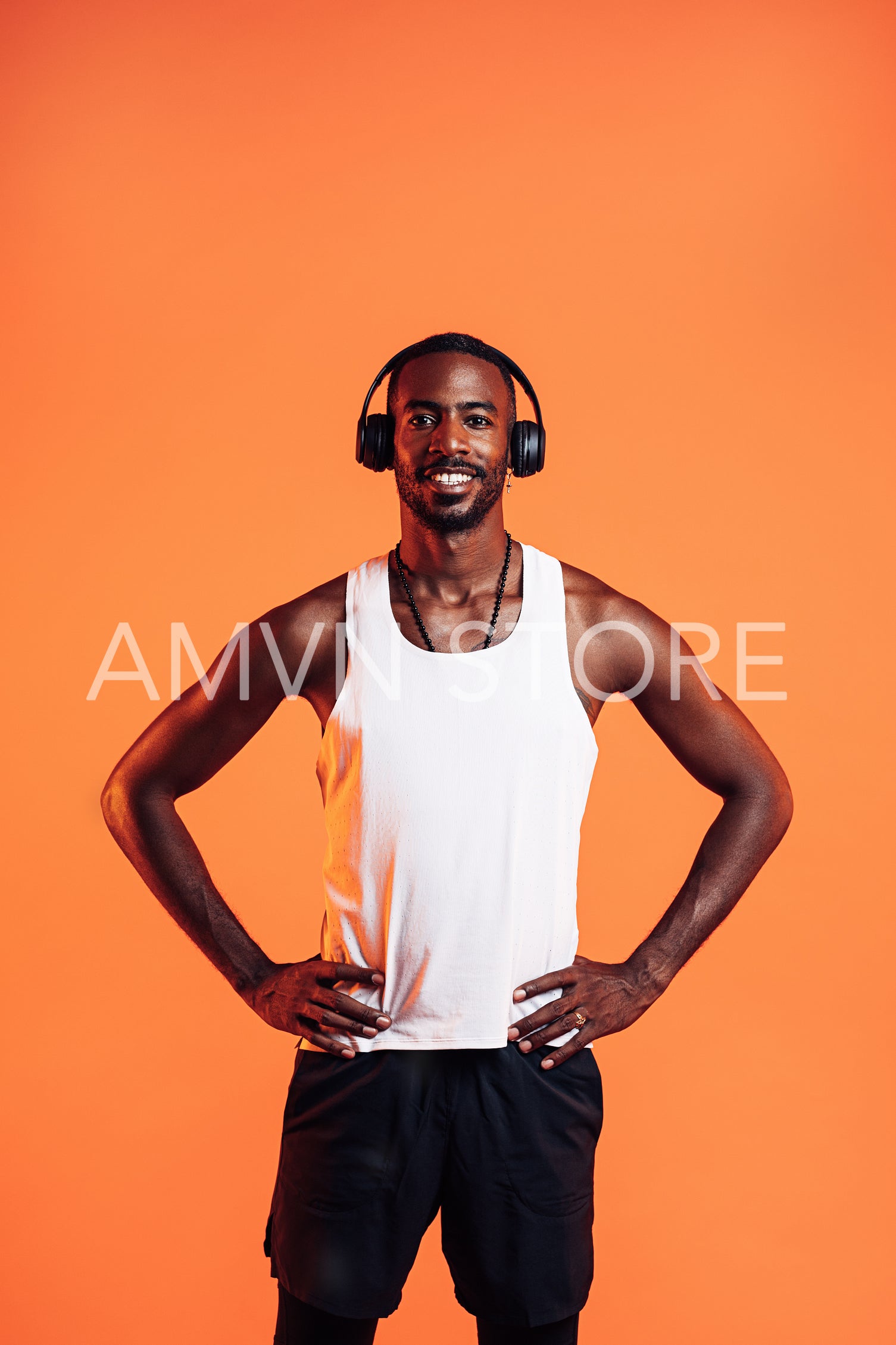 Happy guy wearing wireless headphones standing against an orange background with hands on his waist