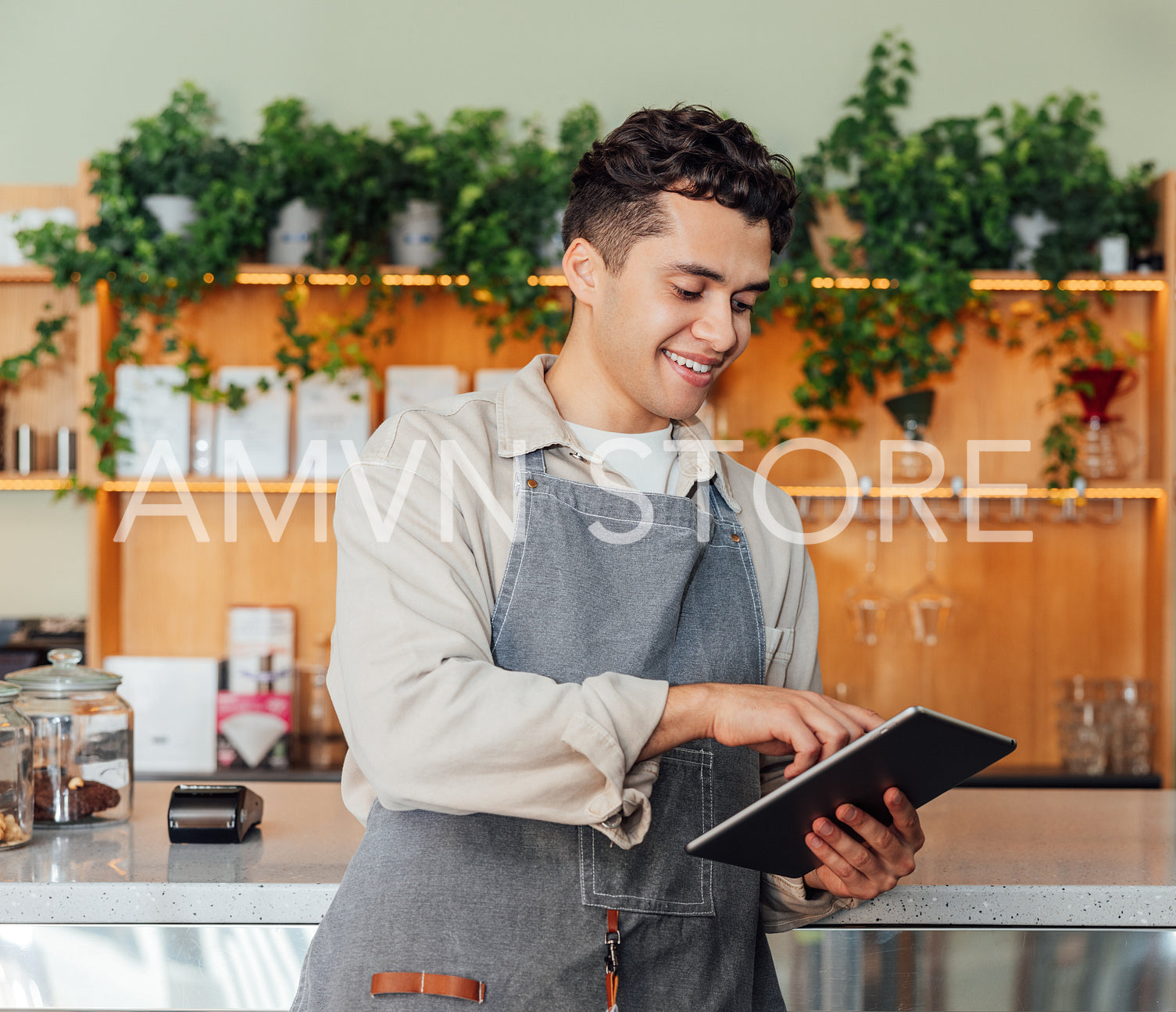 Smiling barista in an apron holding a digital tablet while leaning counter. Coffee shop owner typing on digital tablet.