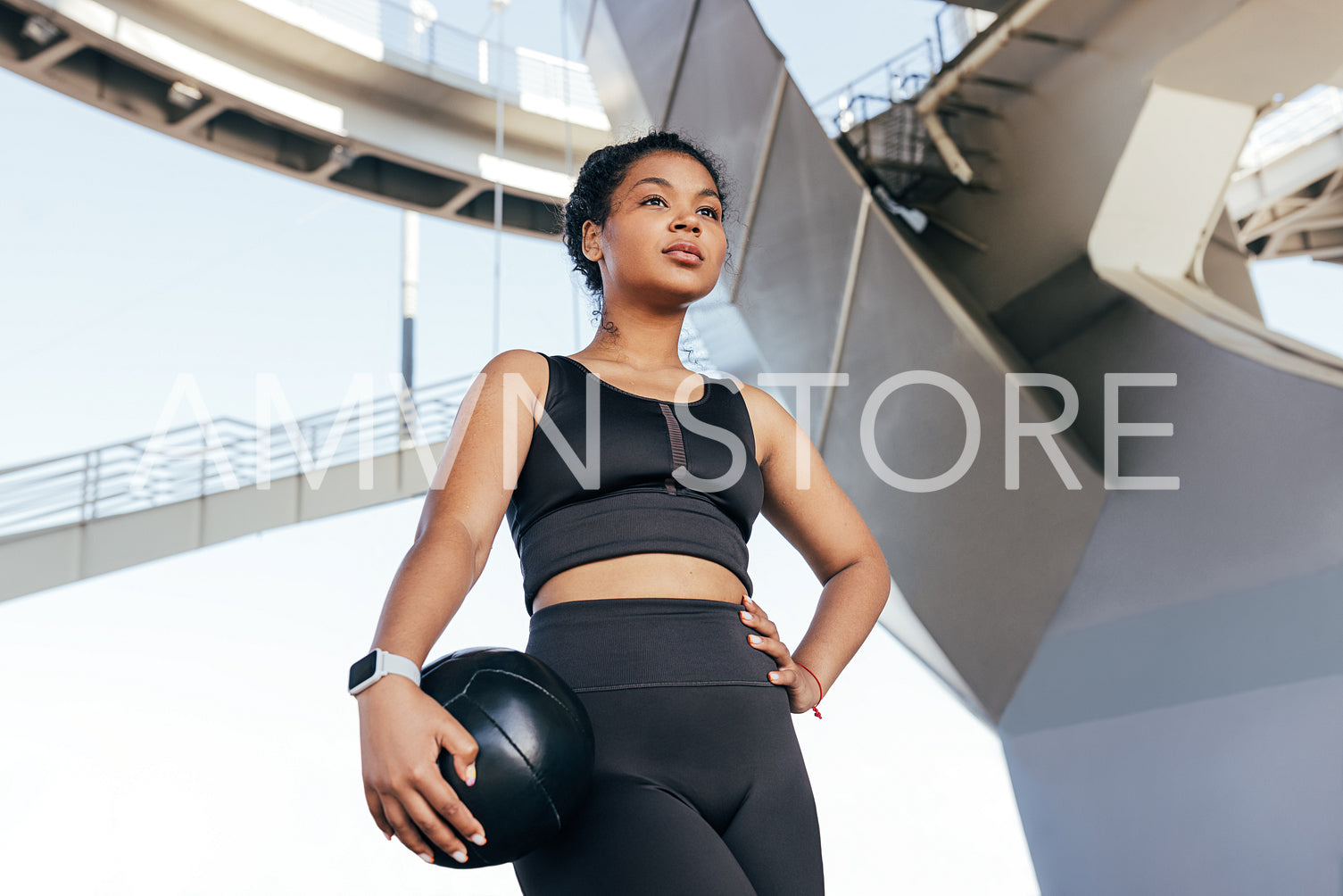 Young slim woman with curly hair in black fitness attire holding a medicine ball standing outdoors