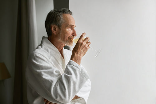 Side view of an elderly man drinking wine while looking at window in living room