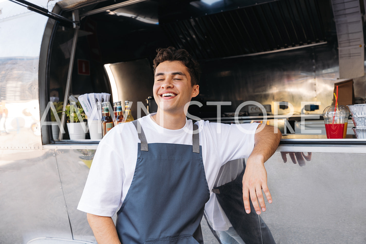 Cheerful food truck owner leaning counter. Young guy working as a chef in a food truck standing with closed eyes and smiling.