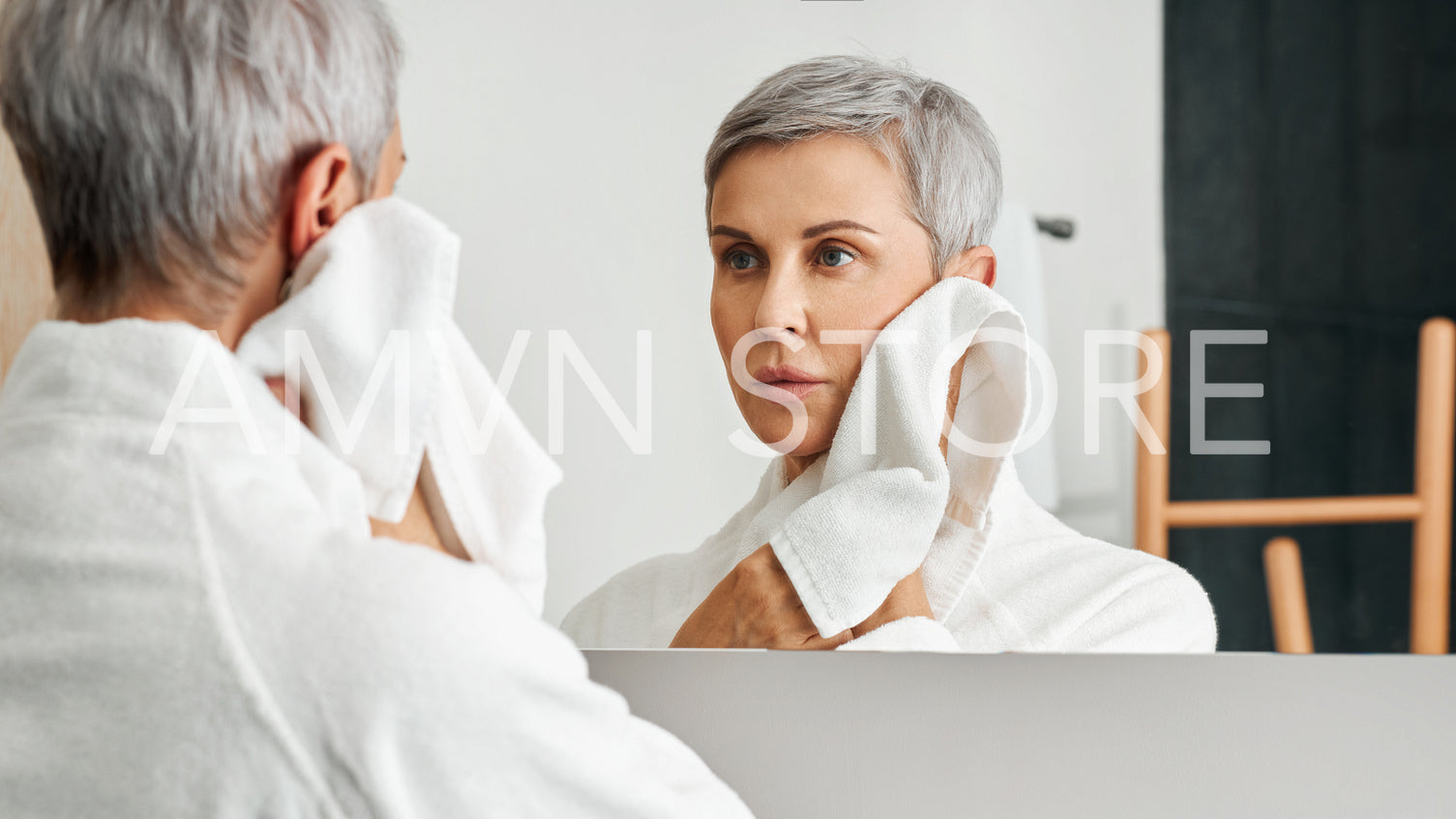 Mature woman with short grey hair looking at her reflection in bathroom mirror while wiping cheek