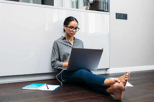 Young businesswoman using laptop sitting on the kitchen floor wearing earphones