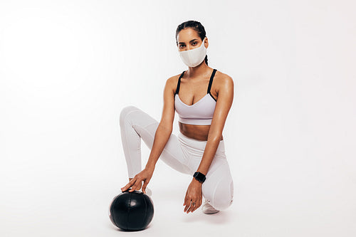 Mixed race woman with face mask sitting in a studio with a medicine ball