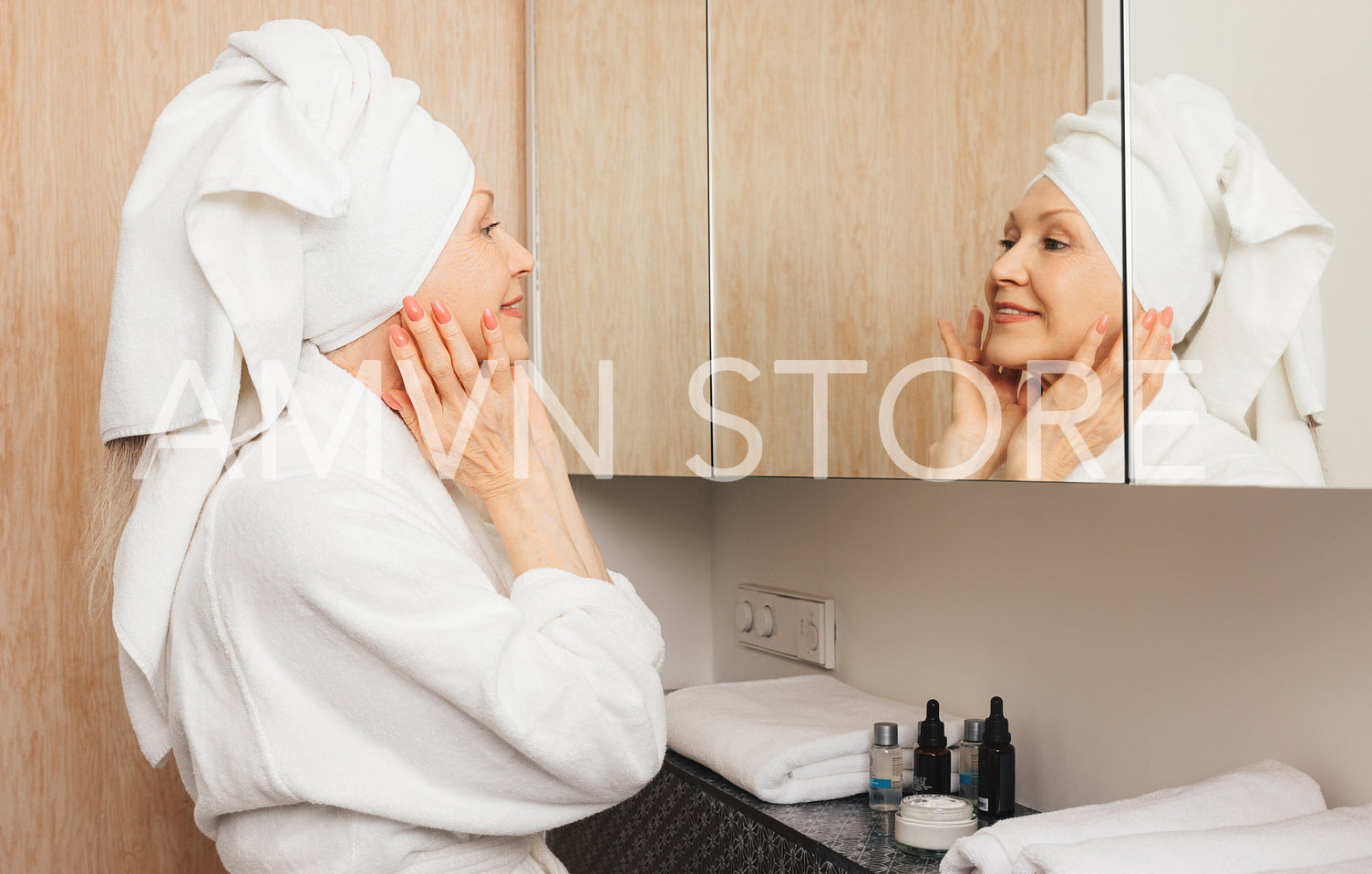 Side view of an aged woman with towel on her head massaging her face while looking at a mirror in the bathroom