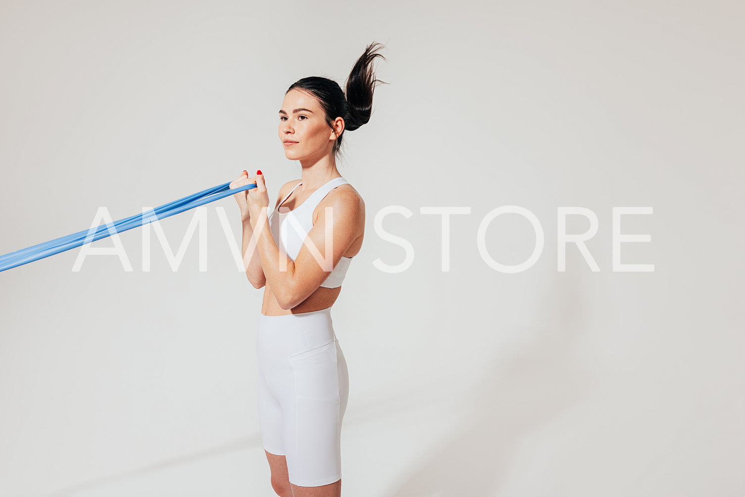 Side view of a slim woman in white fitness attire doing biceps curl with resistance band