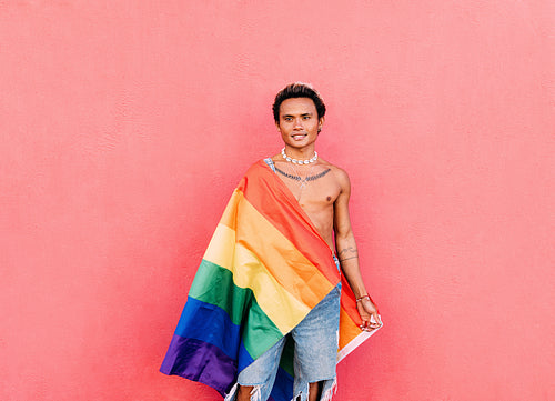 Young handsome man wrapped in rainbow LGBT flag at pink wall