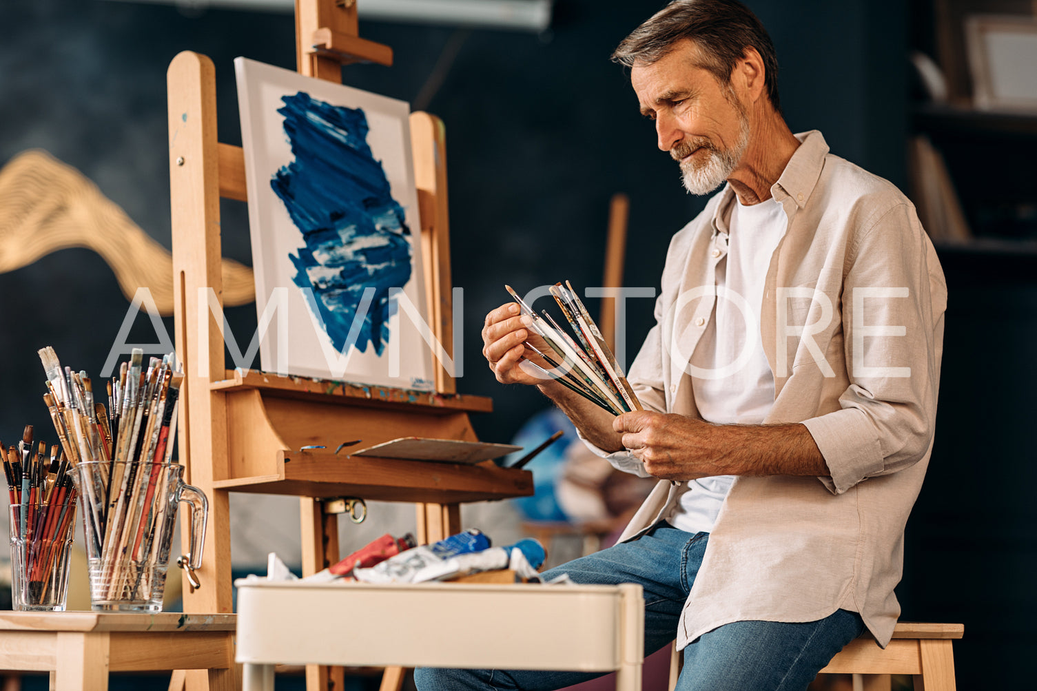 Mature painter holding a bunch of paint brushes sitting in studio	