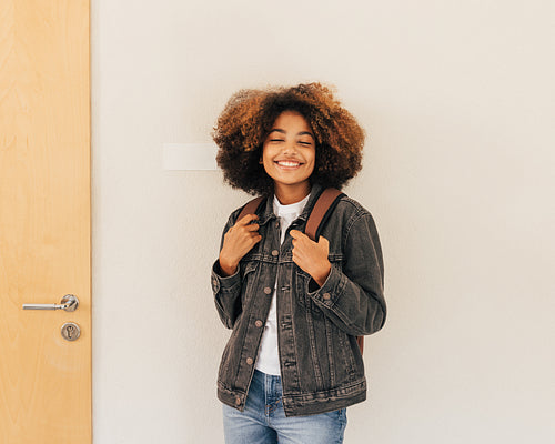 Cheerful girl standing at wall with closed eyes in school. Smiling student with backpack with good mood.