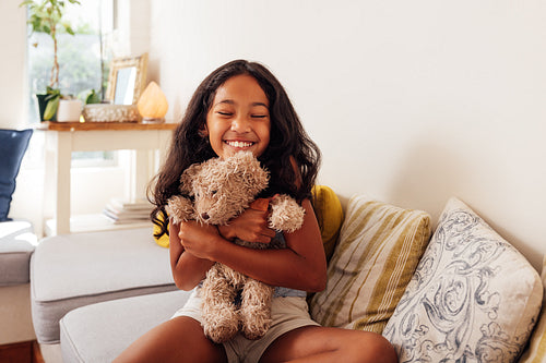 Happy girl with closed eyes hugging her favorite toy while sitting on a sofa in the living room