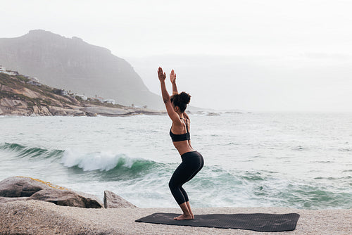 Young woman doing Utkatasana pose by ocean. Slim female in practicing Chair Pose at evening by seaside.