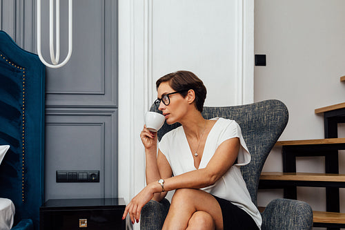 Side view of mid adult woman drinking coffee in a modern apartment. Businesswoman in a hotel room sitting in an armchair and looking away.