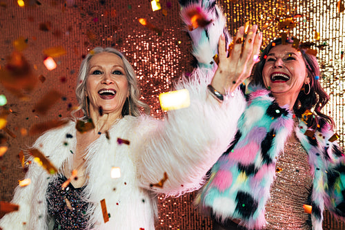 Two cheerful senior females in fur coats dancing under confetti