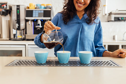 Unrecognizable smiling barista pouring coffee from glass teapot in three blue cups