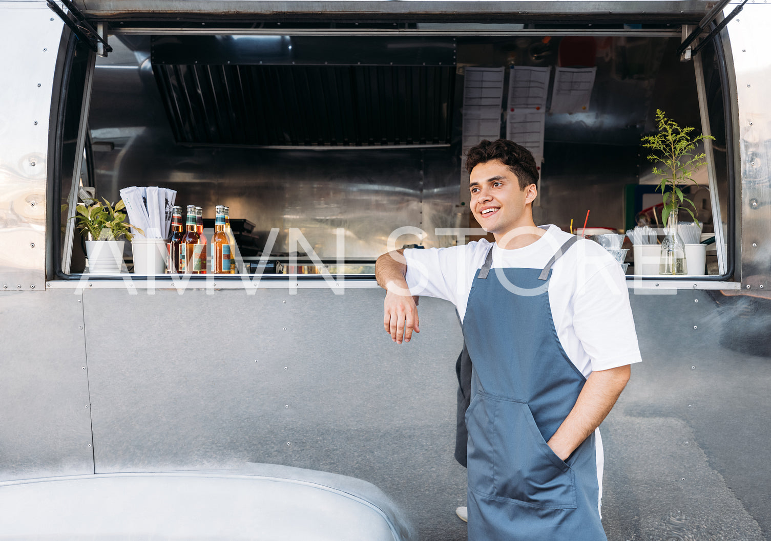 Young business owner in an apron. Salesman leaning on a food truck.