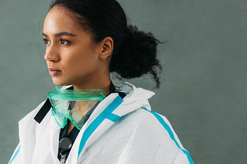 Young serious nurse in a protective suit with glasses standing at wall looking away