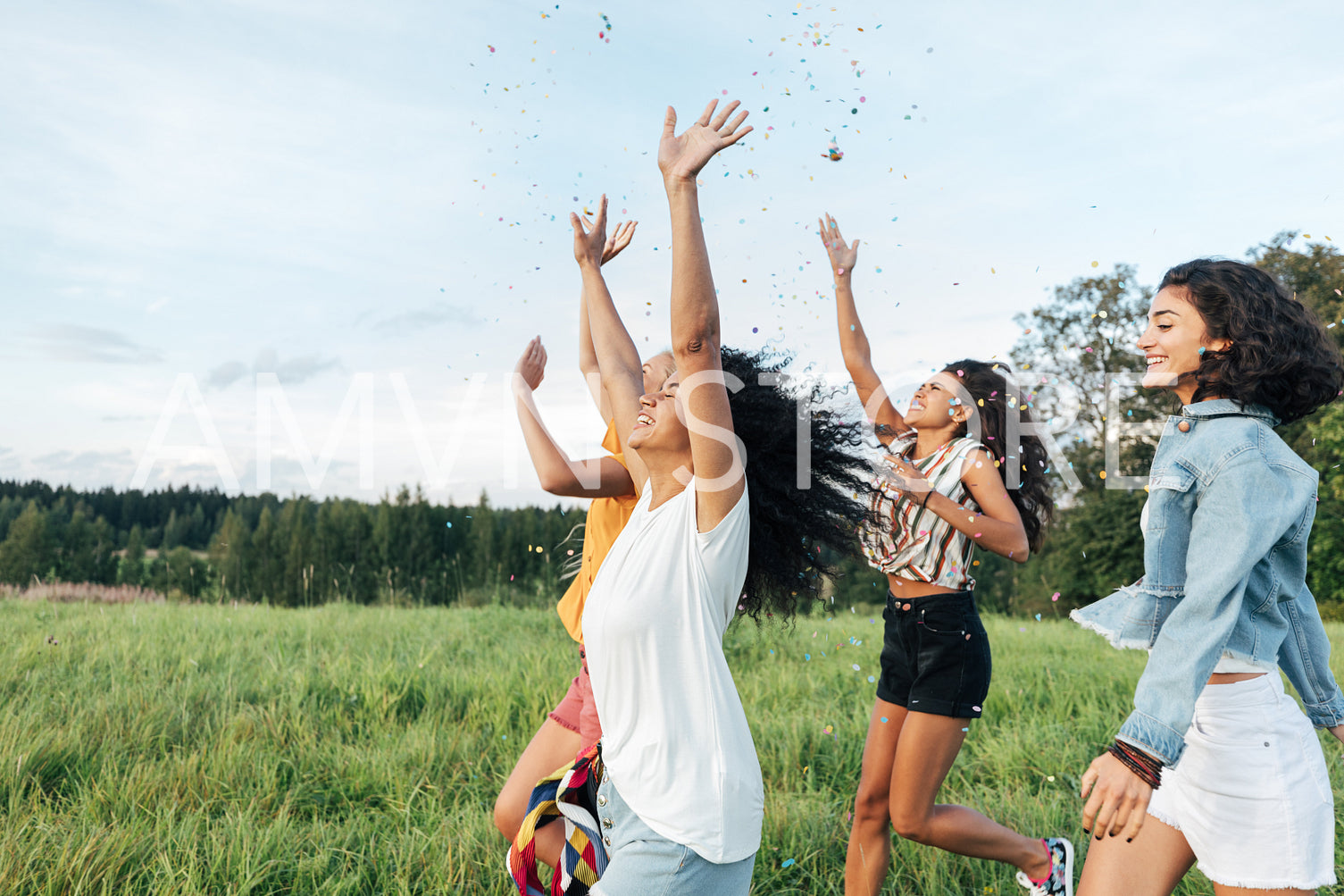 Group of multi-ethnic female friends raised hands up and throwing confetti while running on a field