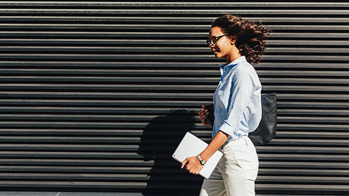 Side view of businesswoman holding a digital tablet, walking outside at wall