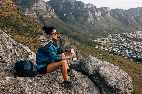 Side view of woman sitting on a rock holding thermos. Female hiker relaxing while sitting on top of a mountain.