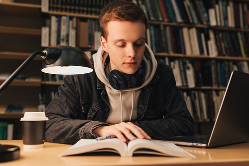 Handsome guy with ginger hair reading book late evening in library sitting at table