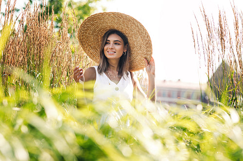 Young happy woman in big straw hat standing on the field and looking away. Stylish female walking outdoors.