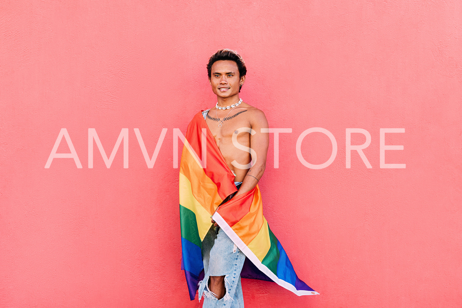 Young stylish man covered by rainbow LGBT flag leaning pink wall