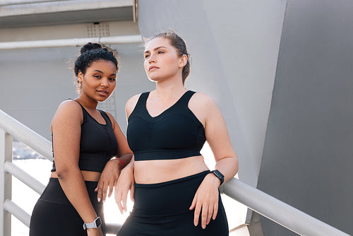 Two plus-size females in black fitness wear posing together after a workout. Young female in fitness attire looking at the camera while standing outdoors with her friend.