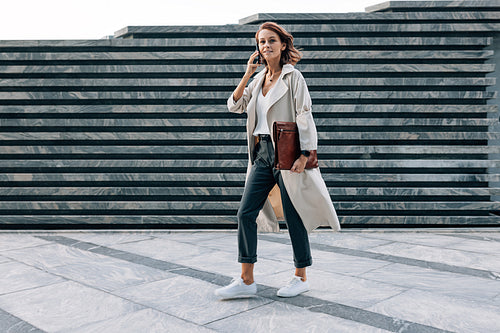 Full length of a walking middle-aged businesswoman with a folder talking on a mobile phone