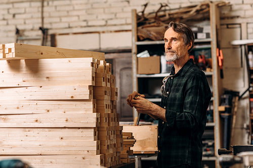 Senior man standing near a bunch wooden parts of furniture in a workshop and looking away