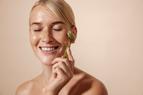 Smiling woman with freckles using jade stone roller against pastel background