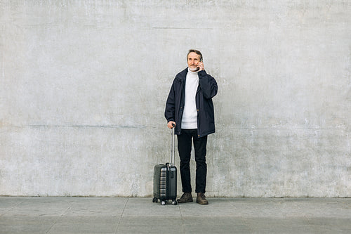 Senior man with suitcase standing outdoors and talking on smartphone near a wall